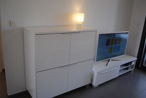 A modern living room with a flat-screen TV on a white entertainment unit, a small table with a lamp, and a wall-mounted light. - Groundfloor Oasis Beach II La Zenia (Orihuela)