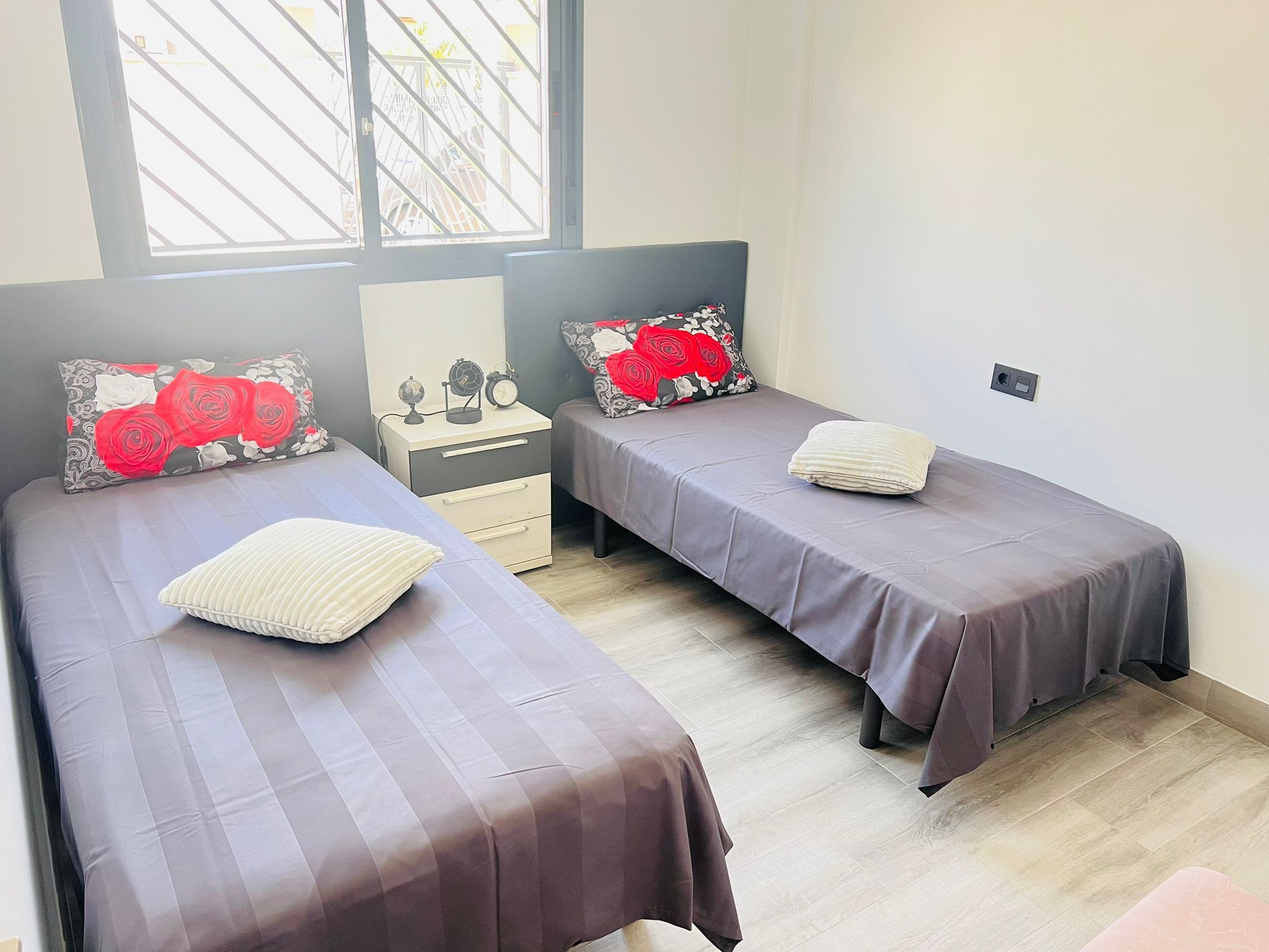 Un dormitorio con dos camas, cada una con una colcha gris y un cojín estampado con motivos florales rojos. Hay una mesita de noche con un globo terráqueo y un despertador.