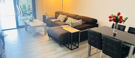 A modern living room with a sofa, coffee table, and dining table. There is a wall art piece and a vase with flowers.