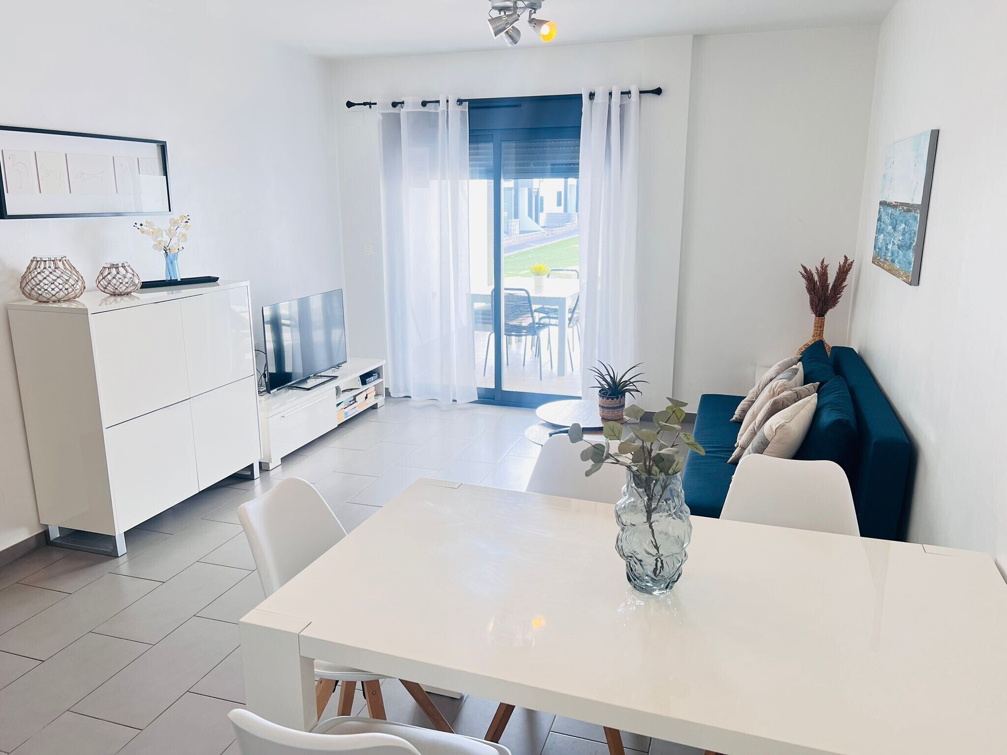 Un salon moderne et bien éclairé, avec une table à manger, un canapé blanc et un téléviseur posé sur un meuble blanc.