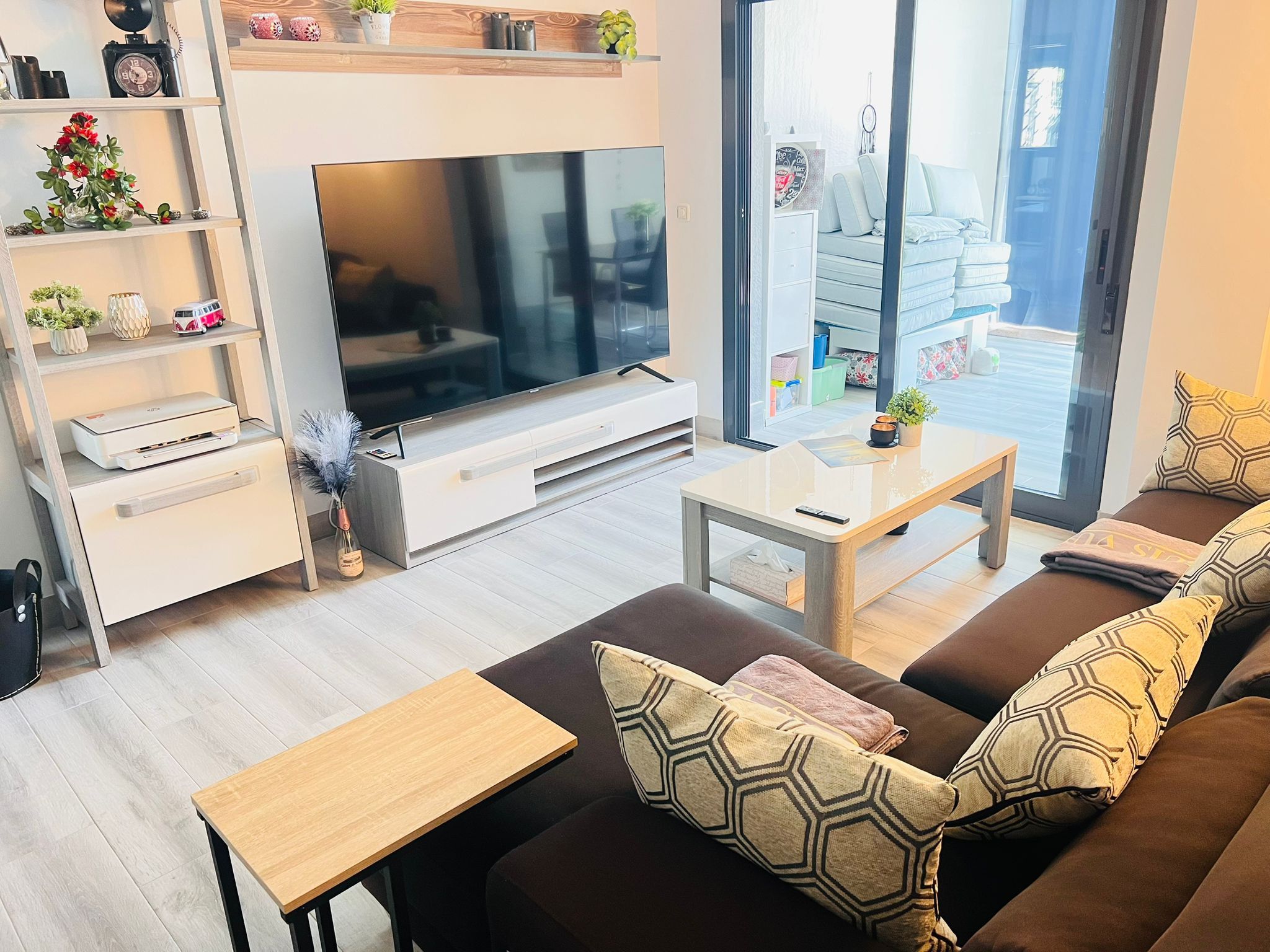 A modern living room with a large flat-screen TV, a sofa with patterned cushions, a coffee table, and a bookshelf with decorative items.