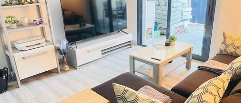 A modern living room with a large flat-screen TV, a sofa with patterned cushions, a coffee table, and a bookshelf with decorative items.