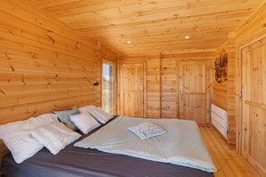 A wooden cabin bedroom with a bed, pillows, a window, and a door. - Holiday Home 'Aurélie' with Wi-Fi (Lavalade)
