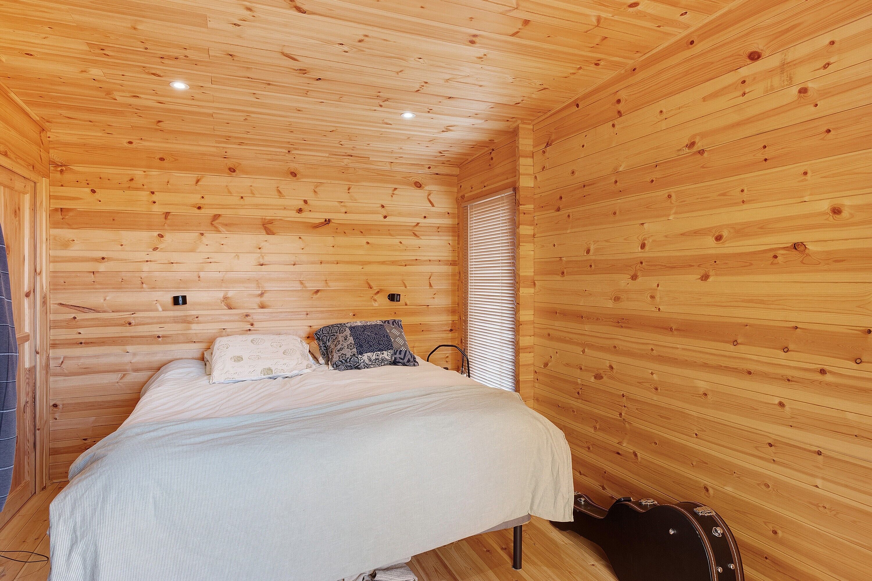 A wooden room with a bed, a window with blinds, and a guitar case.