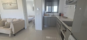 A modern living room with a sofa, a dining area with a table and chairs, a kitchen with a sink and appliances, and a window with blinds. - Apartment Zenia Beach La Zenia (Orihuela)