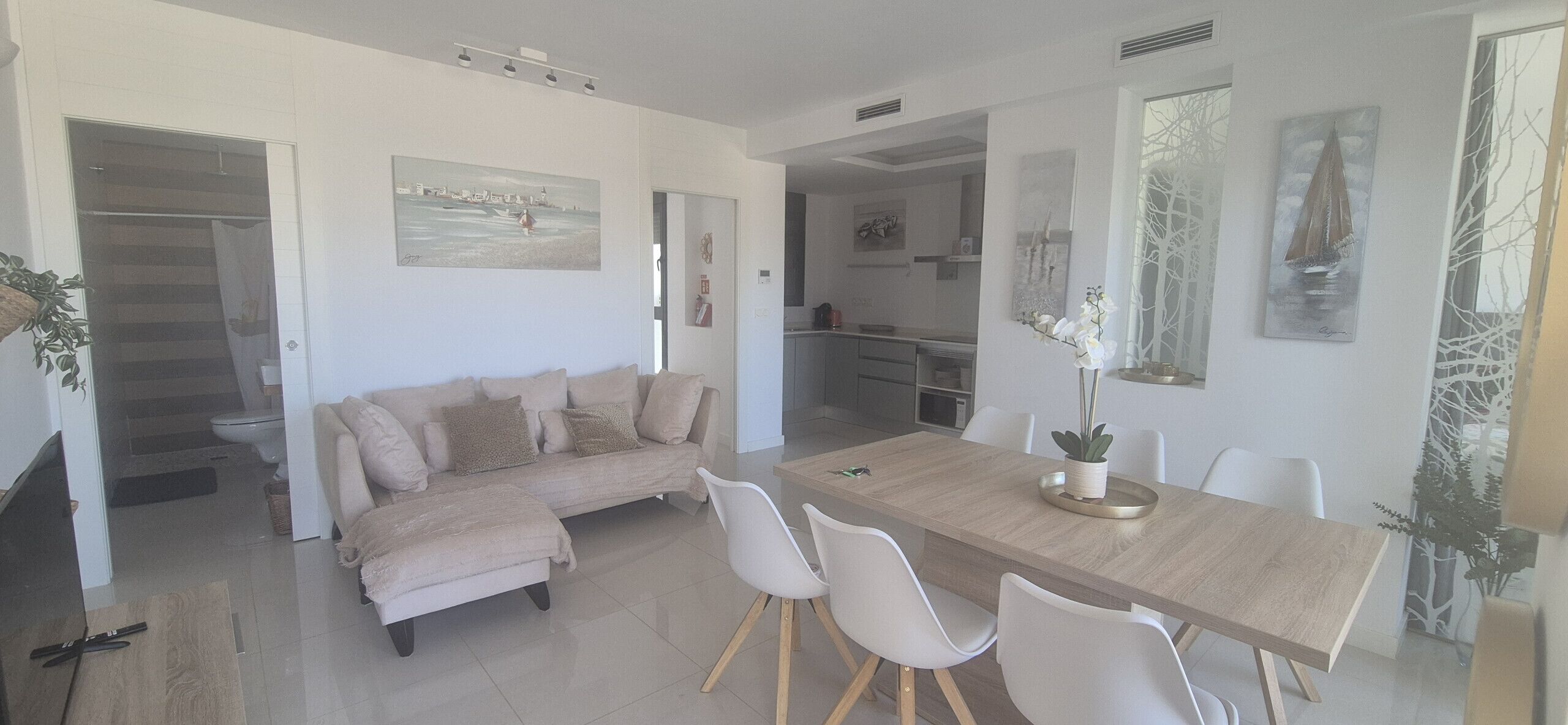 A modern living room with a beige sectional sofa, a wooden dining table with white chairs, and a kitchen in the background.