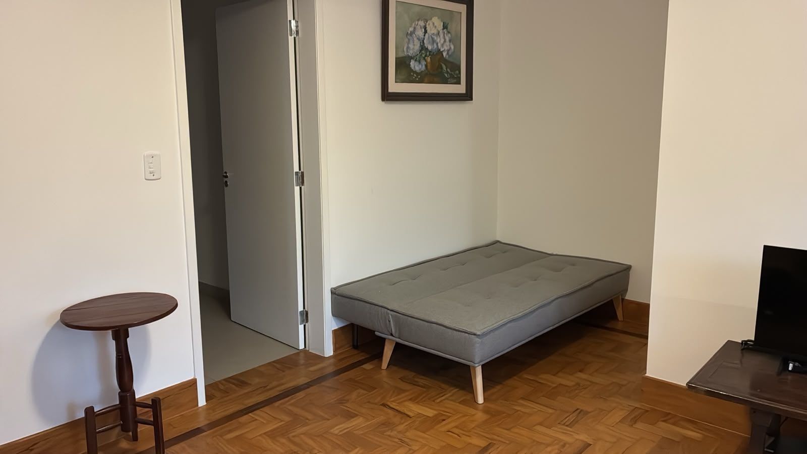Una habitación pequeña con un colchón gris, piso de madera, una mesita redonda y un televisor sobre un soporte.