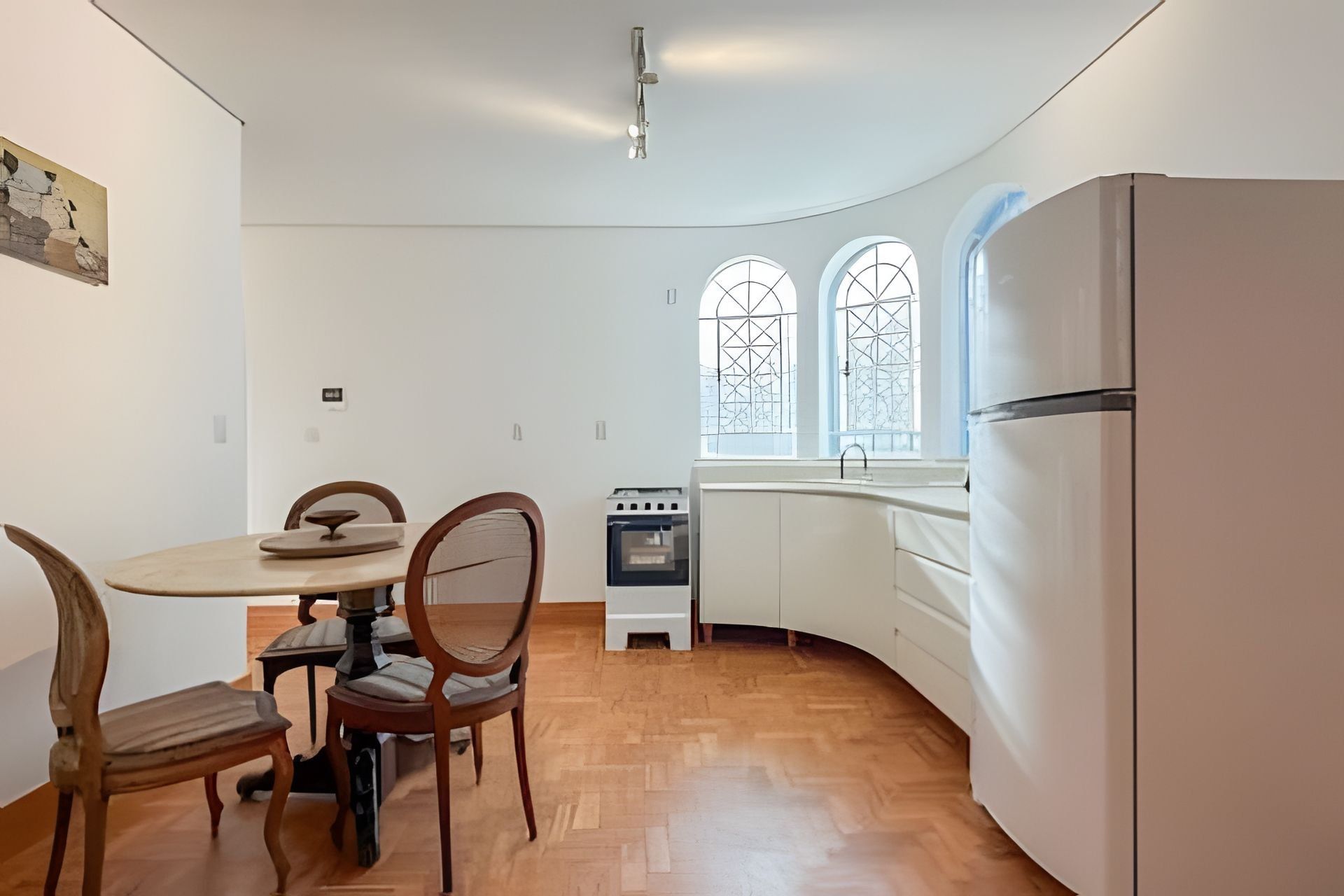 A compact kitchen with a round dining table, wooden chairs, a built-in oven, and a refrigerator.