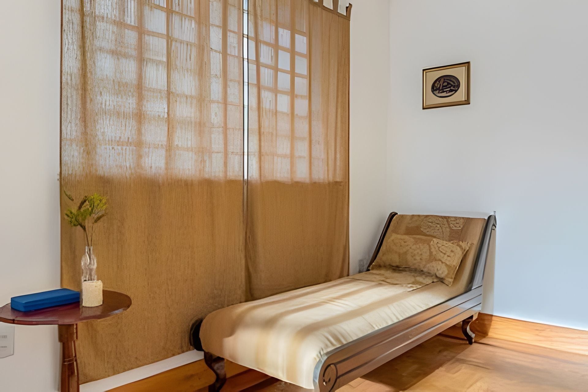A single bed with a wooden frame, a small bedside table with a vase and a blue object, and a window with a wooden blind.