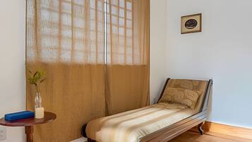 A single bed with a wooden frame, a small bedside table with a vase and a blue object, and a window with a wooden blind.