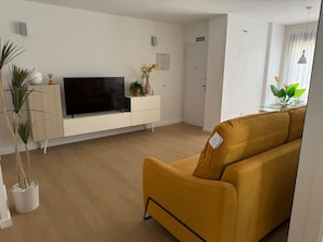 A modern living room with a flat-screen TV on a white console, a mustard-colored sofa, and a potted plant. - Groundfloor Zenia Beach La Zenia (Orihuela)