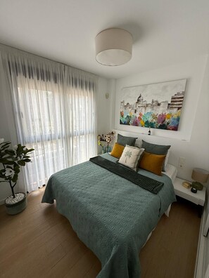 Ein Schlafzimmer mit einem Bett, einem Fenster mit leichtem Vorhang, einem Wandbild und einem Nachttisch mit Lampe.