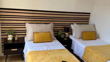 A hotel room with two beds, each with a white bedspread and a yellow throw blanket. There are two bedside tables with decorative items and a wall-mounted headboard.