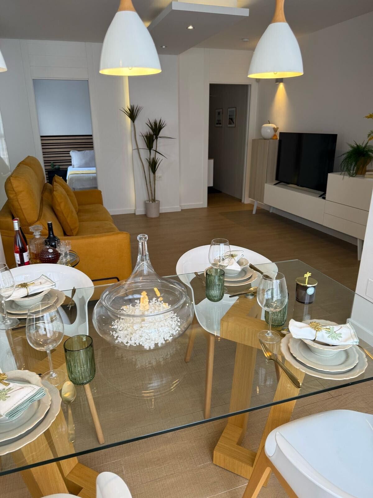 A modern dining area with a glass table set for a meal, surrounded by a living room with a sofa and a TV.