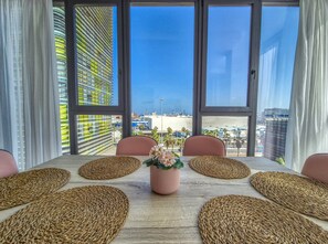 A dining table with woven placemats, a potted plant, and a view of a cityscape through large windows. - House in Gran Canaria Near Playa de Las Canteras (Las Palmas de Gran Canaria)