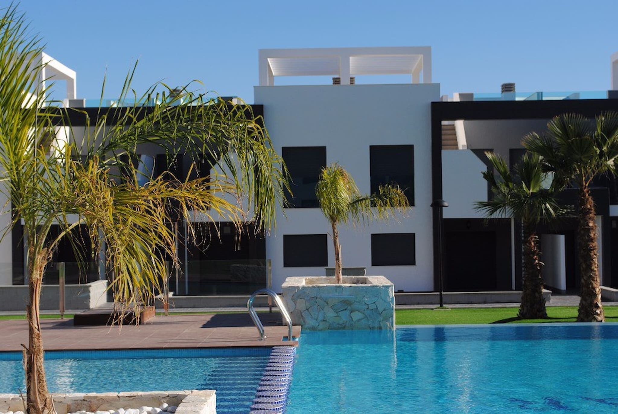 Una piscina amb un edifici modern al fons, palmeres i un cel serè.