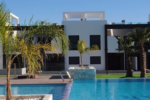 A swimming pool with a modern building in the background, palm trees, and a clear sky. - Groundfloor Oasis Beach I La Zenia (Orihuela)