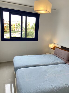 Ein Schlafzimmer mit zwei Betten, einem Fenster mit Blick auf Gebäude und Palmen sowie einer Nachttischlampe.
