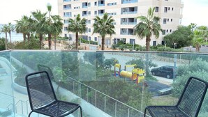 Ein Balkon mit zwei schwarzen Metallstühlen, einer Glasreling und Blick auf einen Spielplatz mit einer gelben Rutsche und einer gelb-roten Spielkonstruktion.