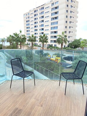 Ein Balkon mit zwei schwarzen Metallstühlen, einer Glasreling und Blick auf einen Spielplatz mit einer gelben Rutsche und einer gelb-roten Spielkonstruktion.