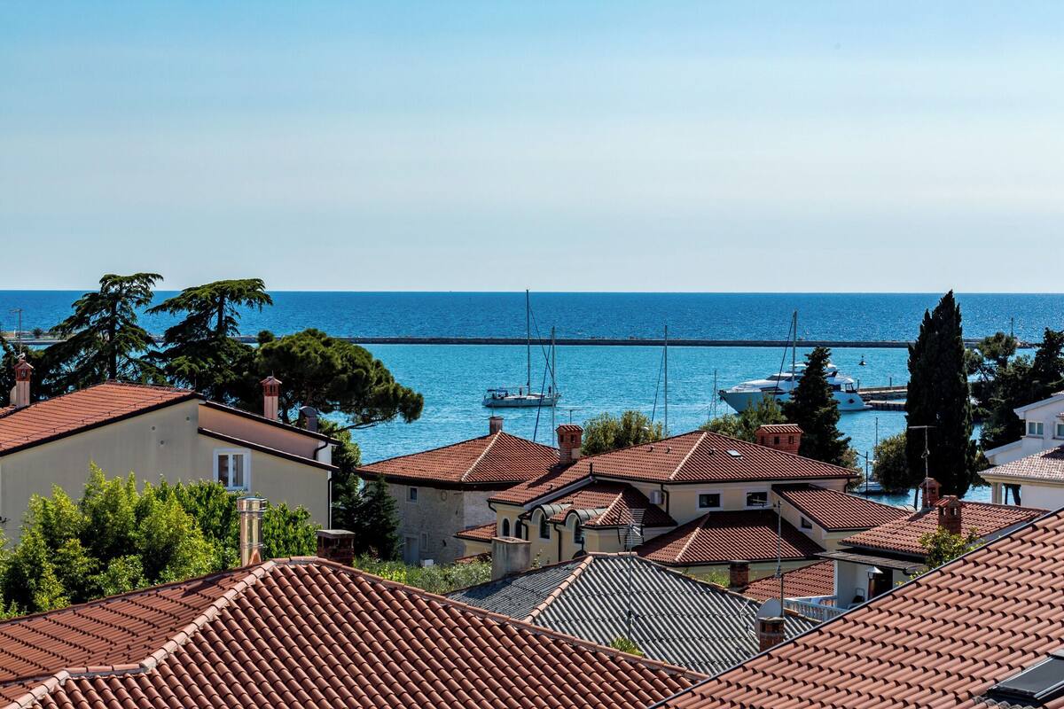 A coastal town with terracotta-roofed buildings, a clear blue sea, and a bridge in the distance.