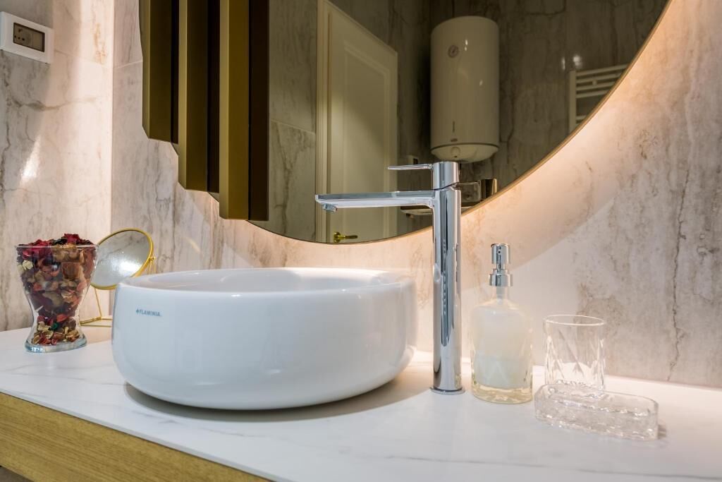 Un baño con lavamanos de mármol blanco, grifo cromado, dispensador de jabón de vidrio y espejo que refleja un perchero y una botella blanca.