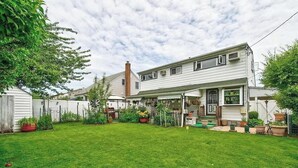 Une maison à deux étages avec un porche couvert, une cour arrière clôturée et diverses plantes en pot.