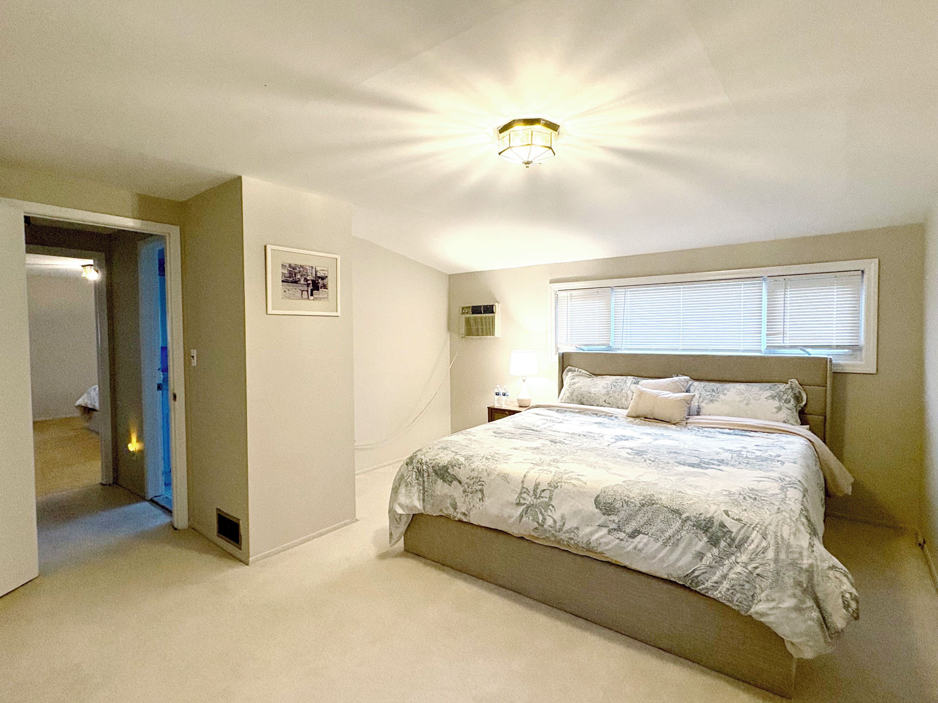 A bedroom with a bed, a nightstand, a window with blinds, a ceiling light, and a door leading to another room.