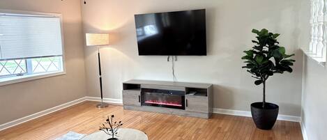 A modern living room with a flat-screen TV, a ceiling fan, a floor lamp, a fireplace, a round coffee table, and a potted plant.