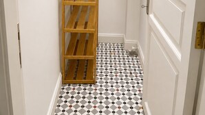 Un couloir étroit avec une étagère en bois, un sac noir sur la deuxième étagère, une fenêtre avec une grille d’aération et une porte blanche avec un cadre blanc.