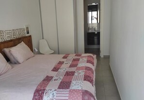 Quarto com cama, cadeira, cabeceira de madeira, parede com espelho e uma kitchenette ao fundo.