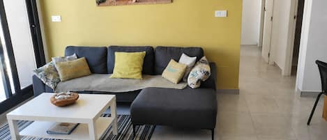 A living room with a sofa, ottoman, and a coffee table. There is a painting on the wall and a window with blinds.