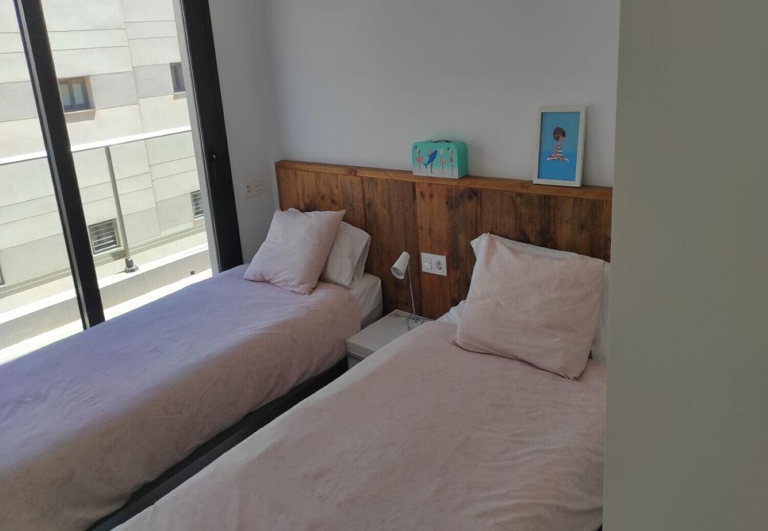 A room with two beds, a wooden headboard, a small table, a lamp, and a window with a view of buildings.