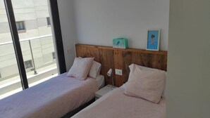 Quarto com duas camas, cabeceira de madeira, uma mesinha, uma luminária e janela com vista para prédios.