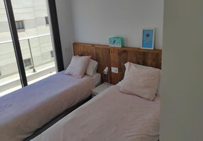 Ein Zimmer mit zwei Betten, einem Holzkopfstück, einem kleinen Tisch, einer Lampe und einem Fenster mit Blick auf Gebäude.