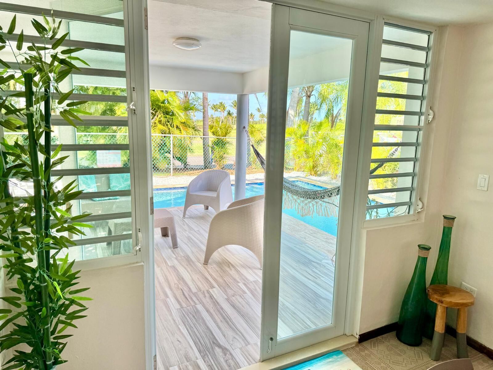 Una habitació amb una porta corredissa de vidre que dona a una zona de piscina, una cadira de vimet blanc i un tamboret de fusta petit.