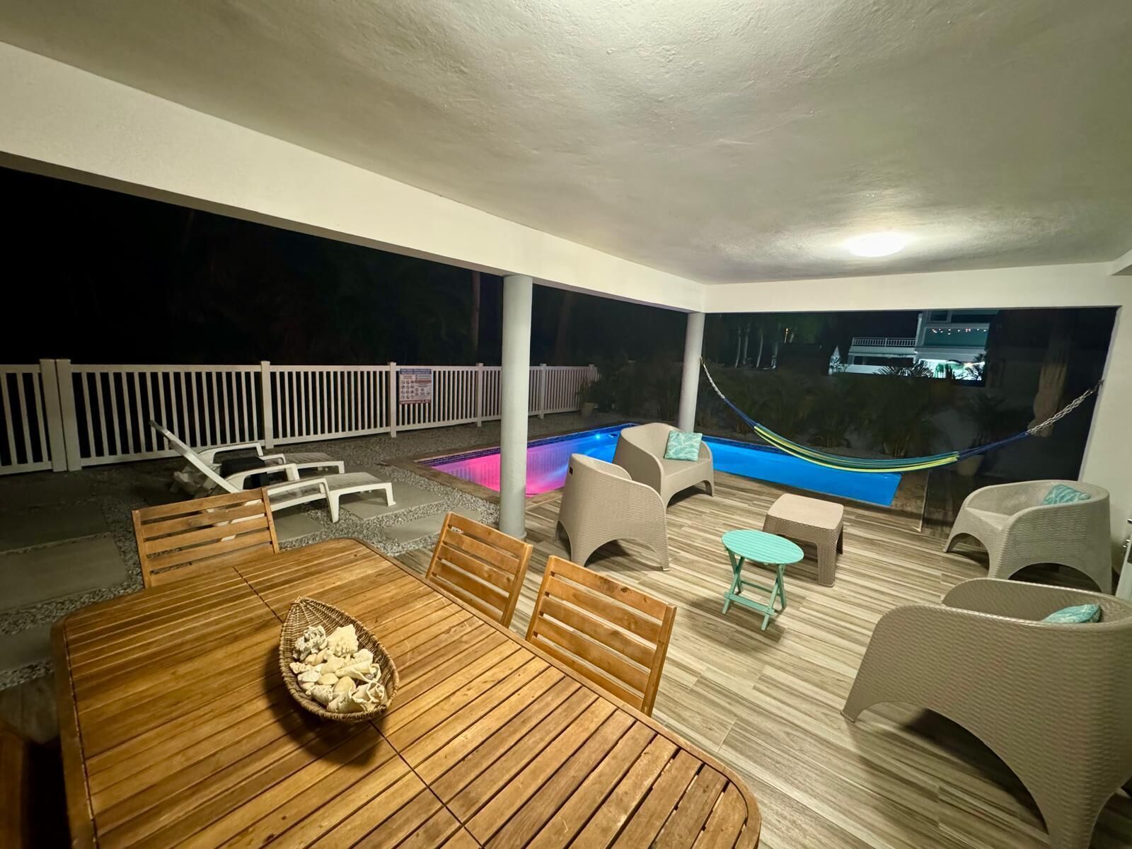 Un patio al aire libre con piscina, mobiliario de madera y cerca blanca.