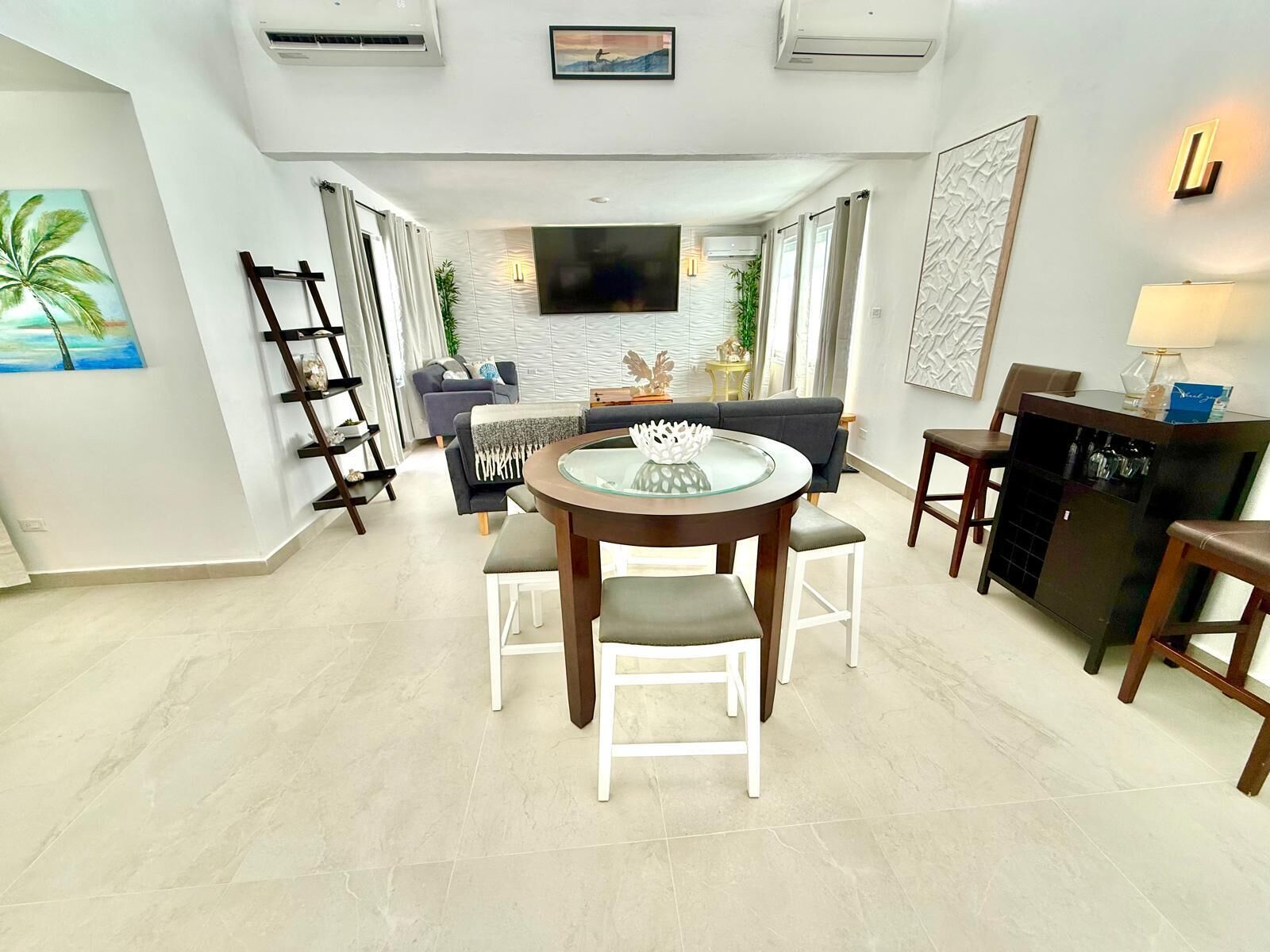 A modern living room with a round glass table, white chairs, a sofa, and a TV mounted on the wall.