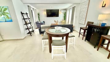 A modern living room with a round glass table, white chairs, a sofa, and a TV mounted on the wall.