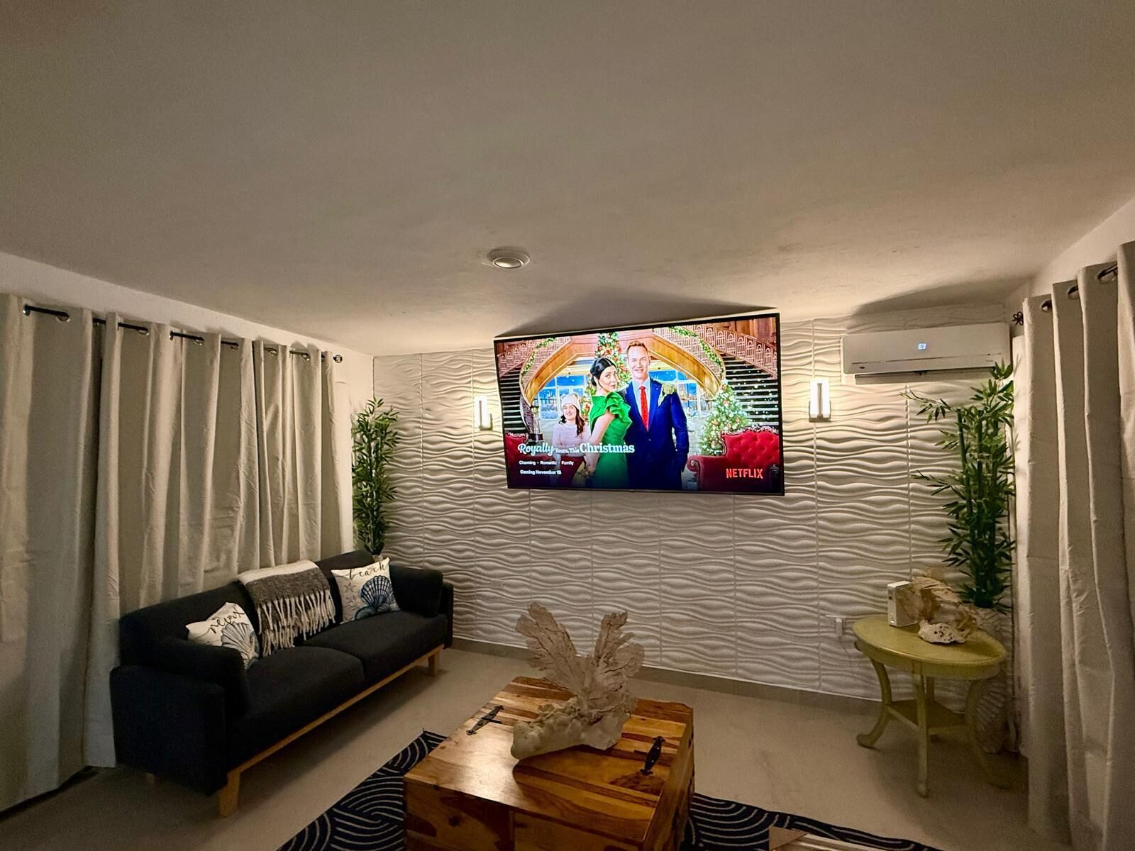 Un salón moderno con un televisor de pantalla plana montado en la pared, un sofá negro, una mesa de centro de madera y una mesita auxiliar con una planta.