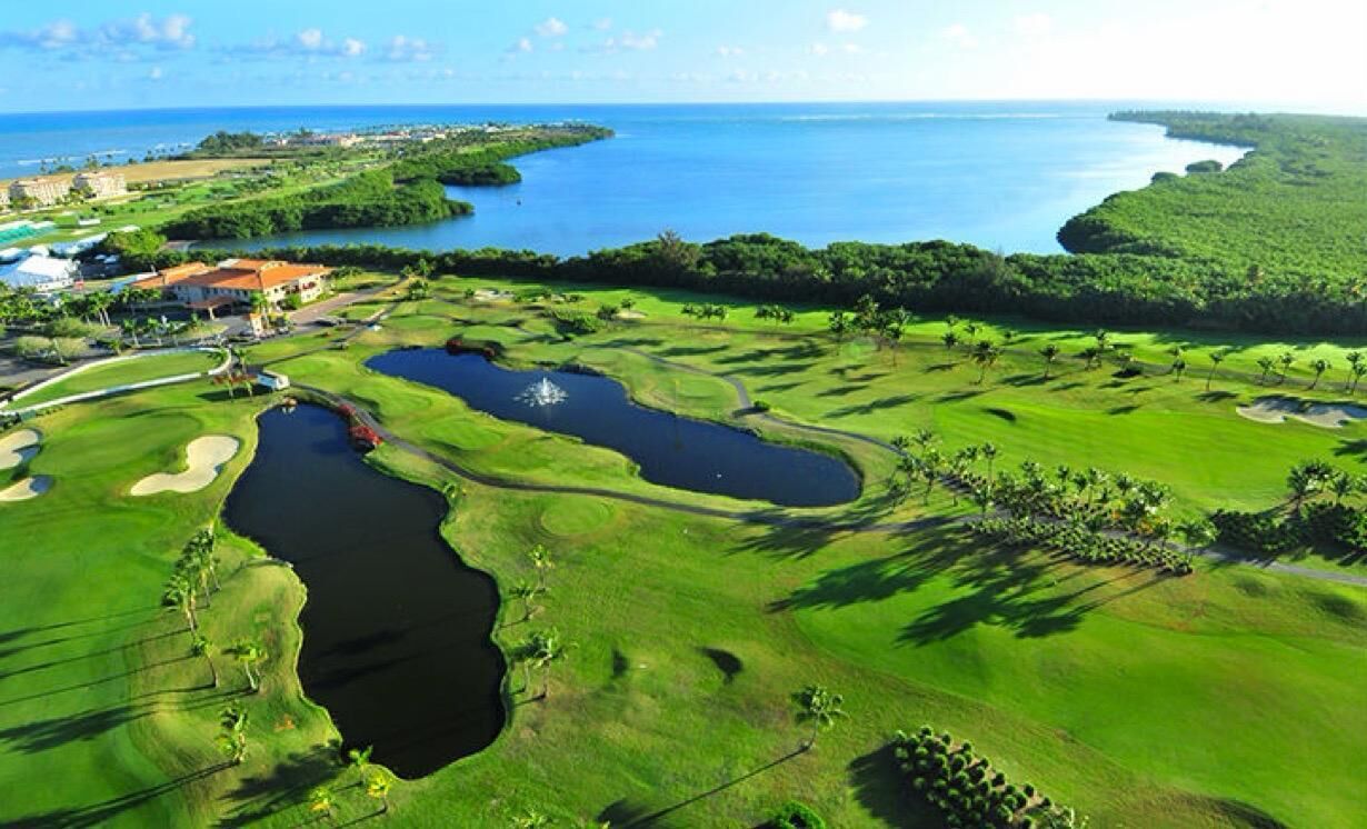 Campo de golf con varios obstáculos de agua, un club de campo y vistas al océano.