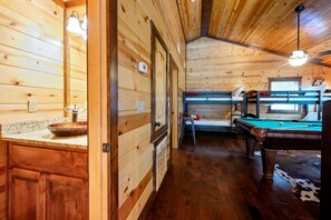 A wooden cabin interior with a pool table, bunk beds, and a kitchen area. - Over Yonder Inn | Hot Tub, Firepit & Pool Table (Broken Bow)