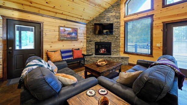 A cozy living room with a stone fireplace, leather sofas, and a large window. - Over Yonder Inn | Hot Tub, Firepit & Pool Table (Broken Bow)