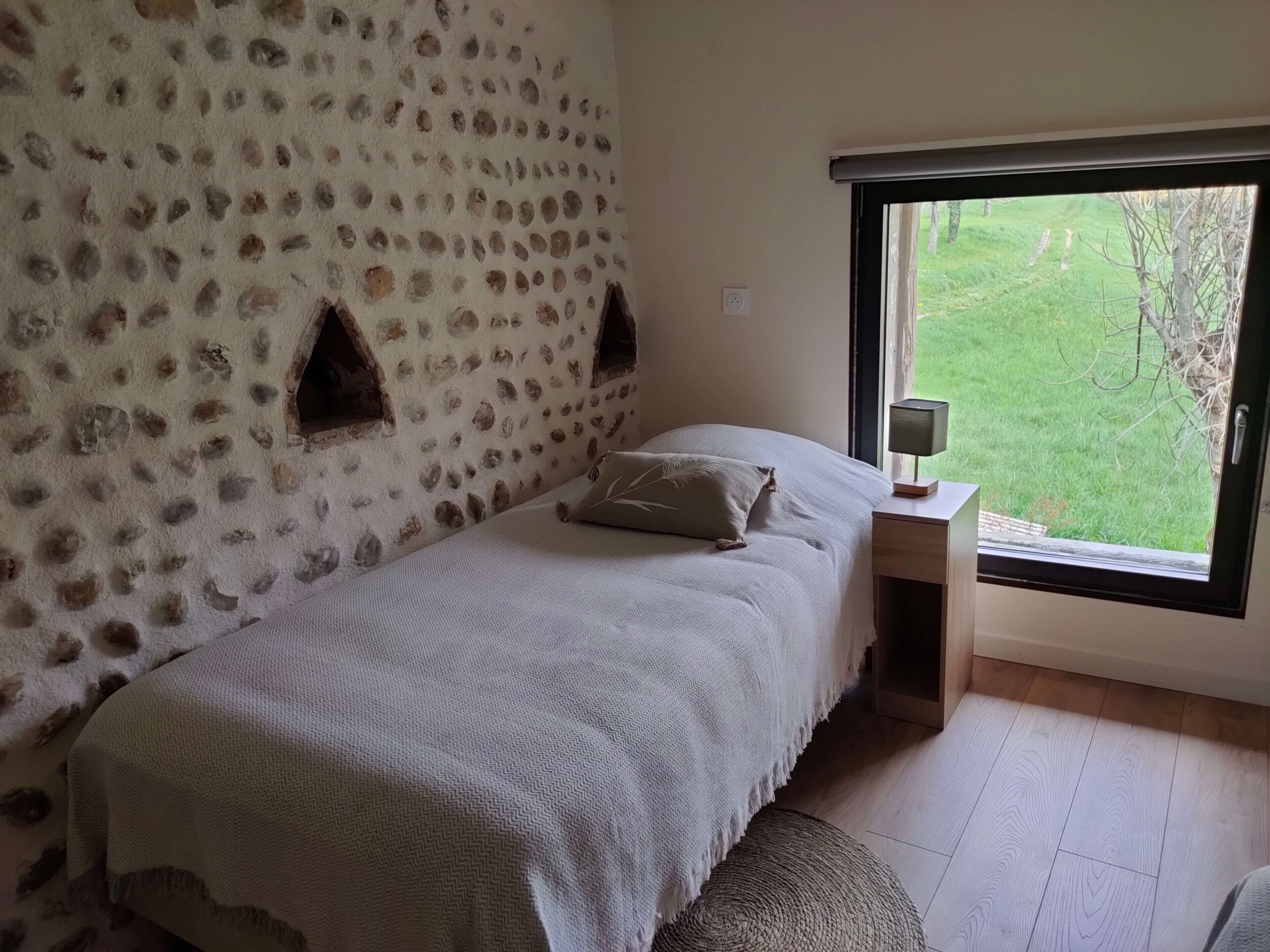 A bedroom with a stone wall, a bed with a white bedspread, a bedside table with a lamp, and a window with a view of greenery.