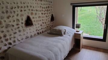 A bedroom with a stone wall, a bed with a white bedspread, a bedside table with a lamp, and a window with a view of greenery.