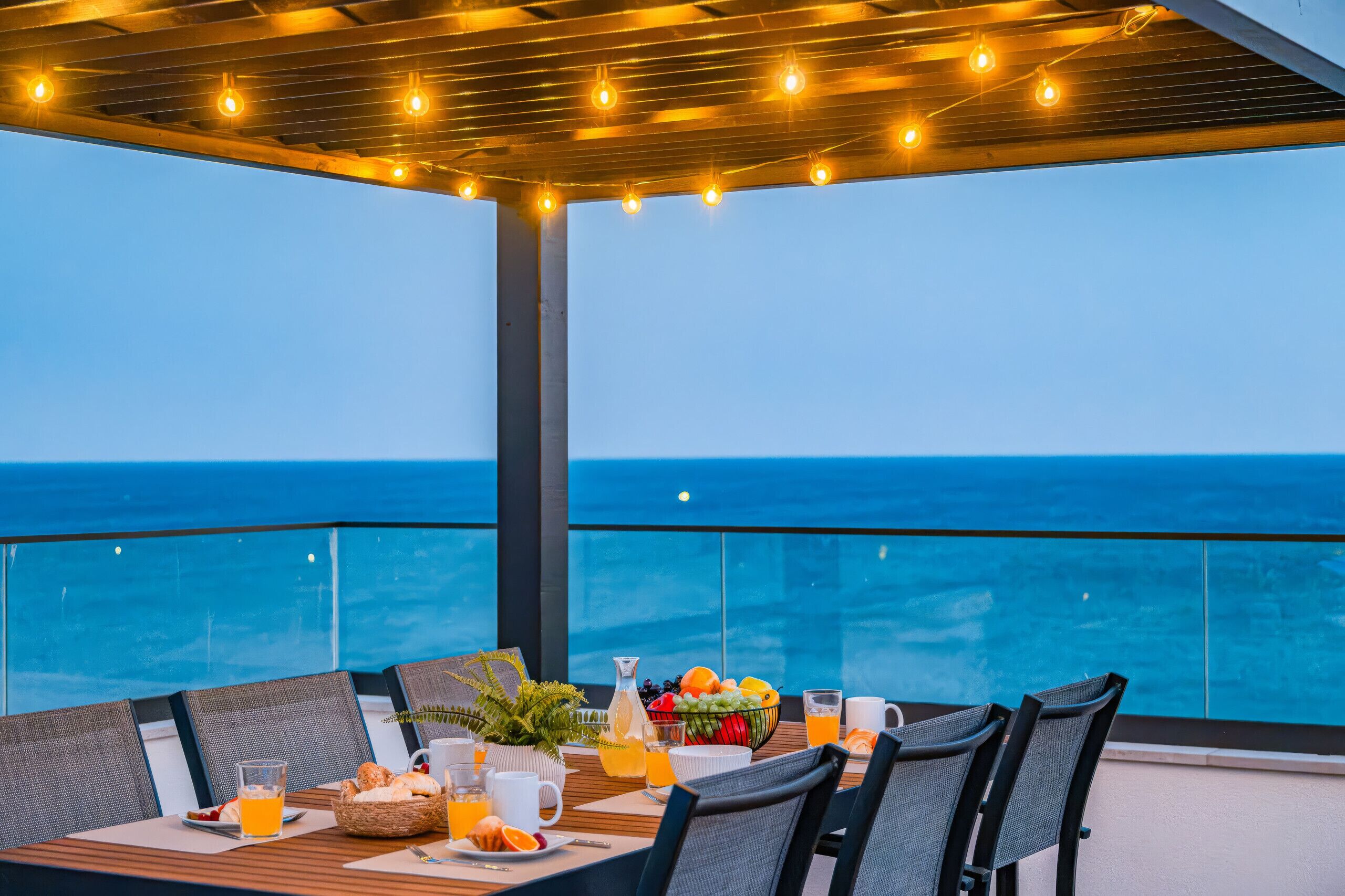 Un espace de restauration en plein air offrant une vue sur l’océan, avec une table dressée pour le petit-déjeuner et une pergola illuminée de guirlandes lumineuses.