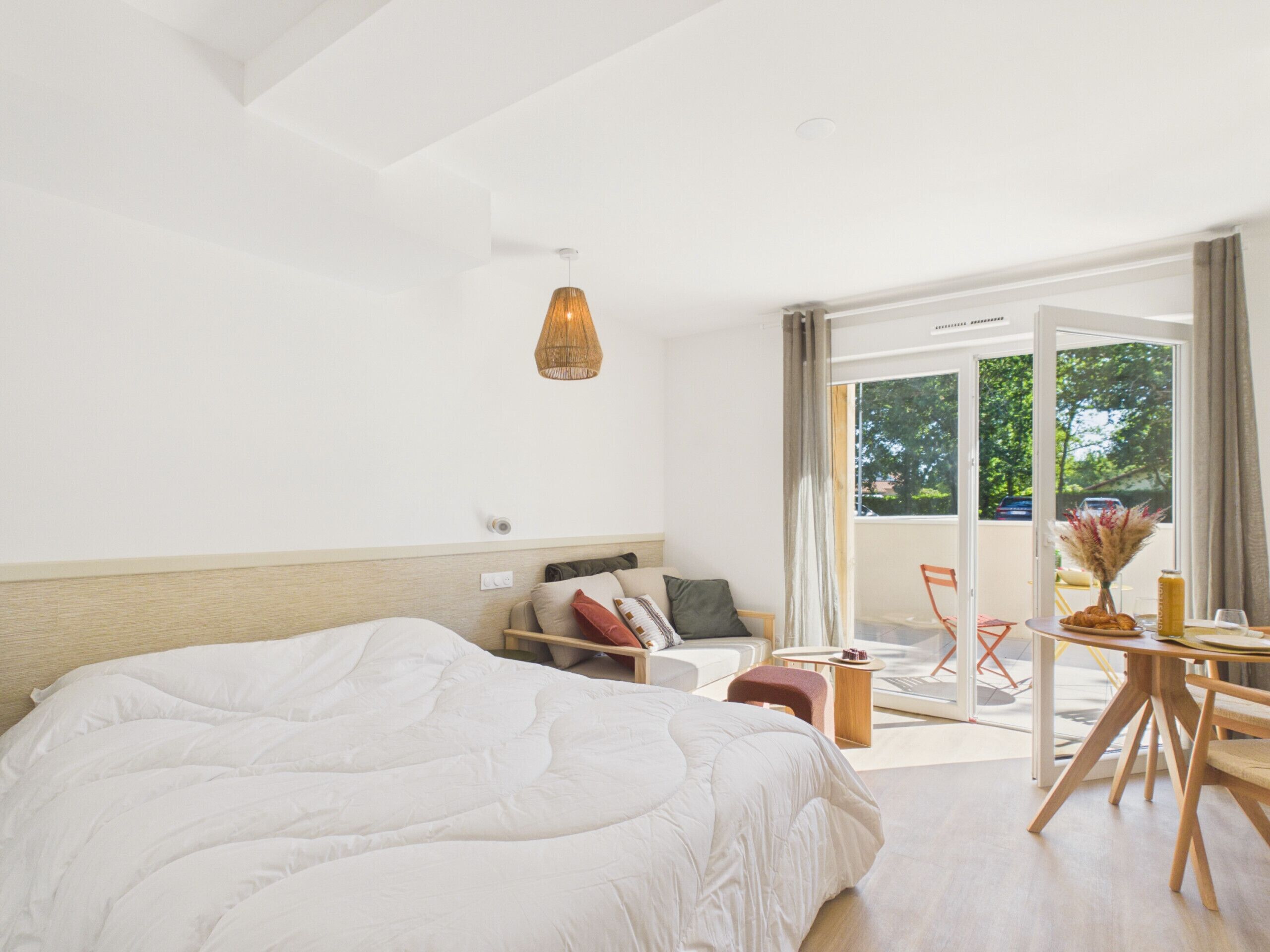 A modern hotel room with a large bed, a sofa, a dining table, and a view of the outdoors.