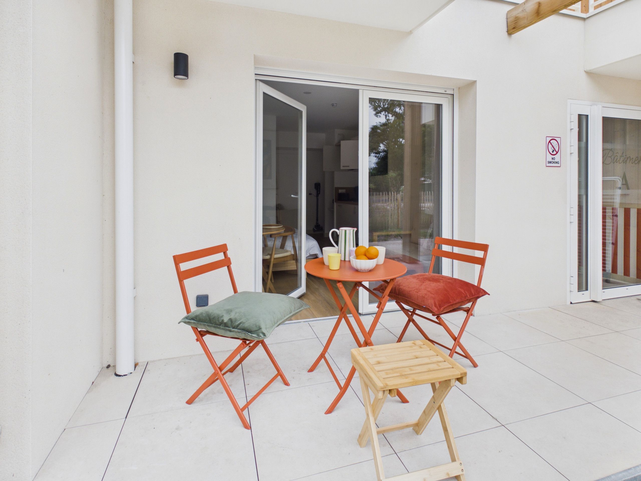 Un espace extérieur aménagé avec une table et deux chaises, un petit tabouret et une porte vitrée donnant sur un espace intérieur.