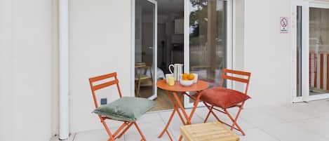 An outdoor seating area with a table and two chairs, a small stool, and a glass door leading to an interior space.