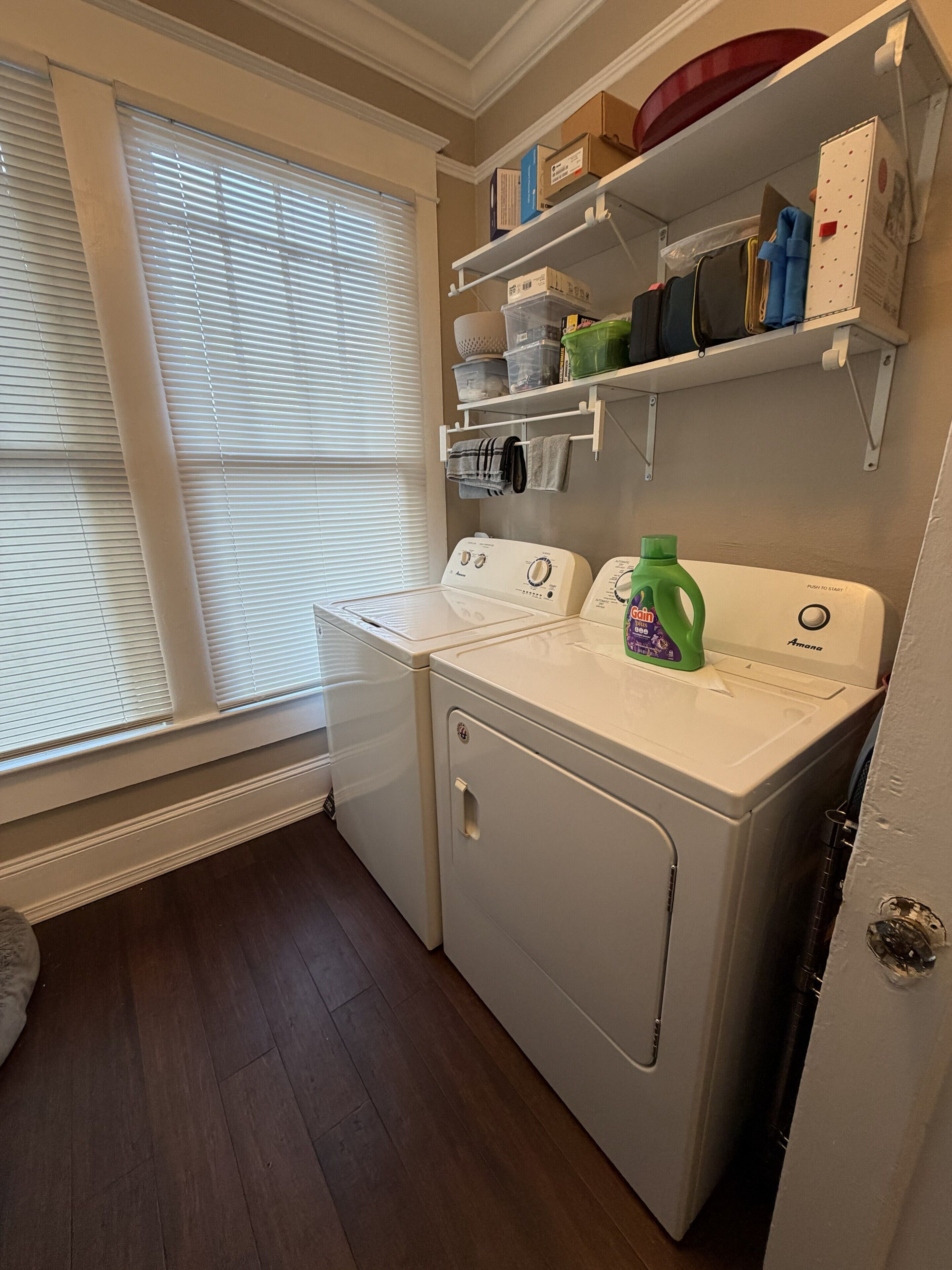 A laundry room with a washer and dryer, shelves with storage boxes, and a window with blinds.